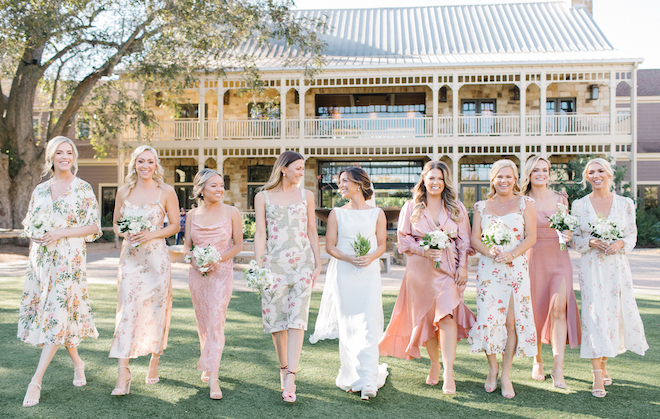 The bride walking with her bridesmaids' who are wearing floral, pink and green dresses before the outdoor autumn wedding.