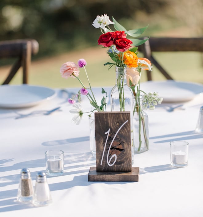 The table number on an outdoor table written on a piece of wood in white letters reading "16" with red, orange, white and pink delicate flowers behind it.
