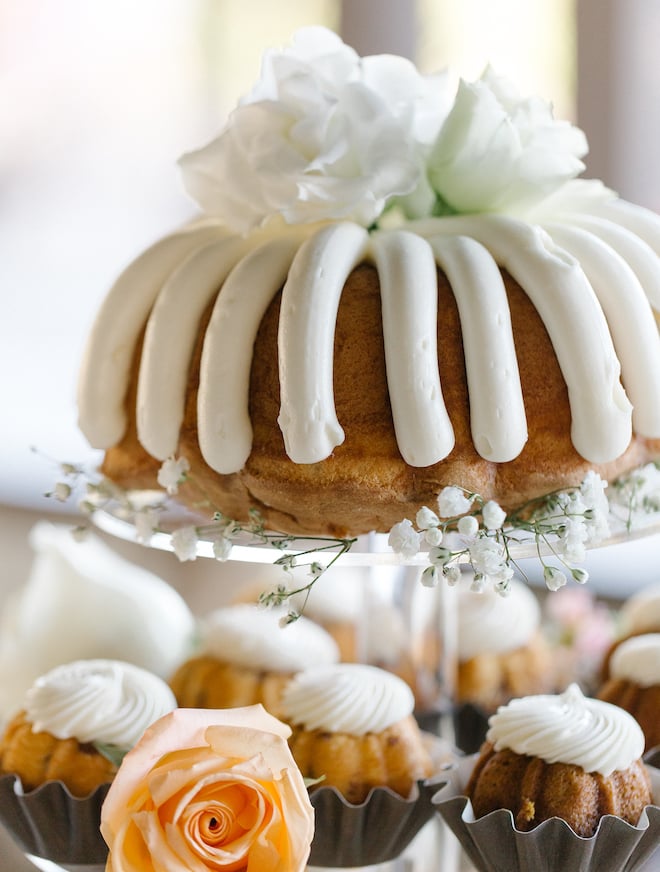 A bundt cake sitting on delicate white flowers with white icing and white flowers topping it, with mini bundt cakes underneath.