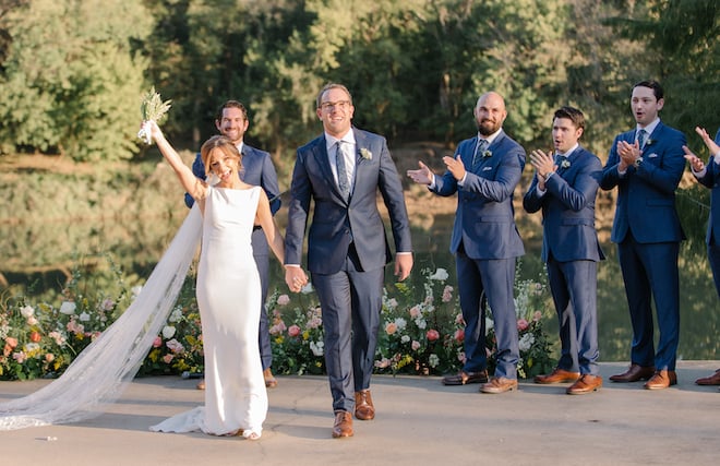The bride cheering as she holds the groom's hand leaving the ceremony, with the groomsmen clapping in the background. 