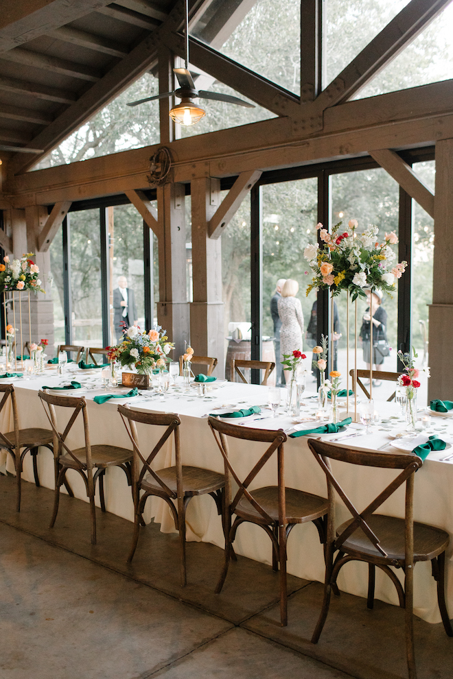 A long reception table decorated with white green and multi-colored florals, next to grand windows in the reception hall at Hyatt Lost Pines for their outdoor autumn wedding.