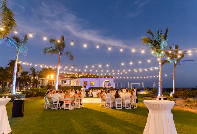 The guests seated at their tables as it gets darker, with string lights crossing the palm trees above them.