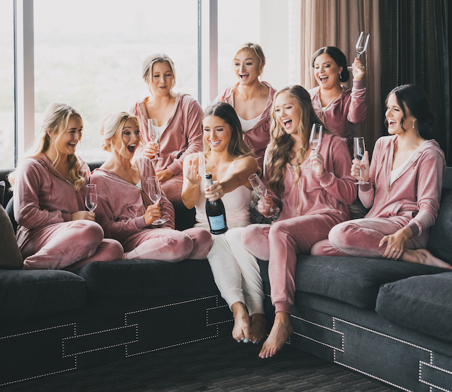 The bride popping champagne with her bridesmaids in their pink sweatsuits.