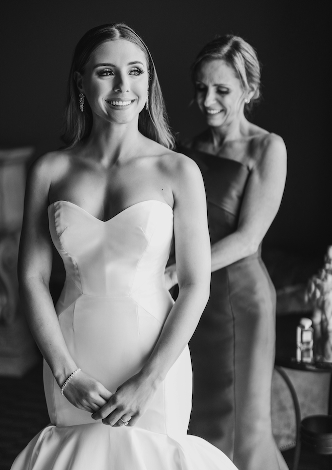 The bride smiling as a woman helps button the back of her glamorous dress.