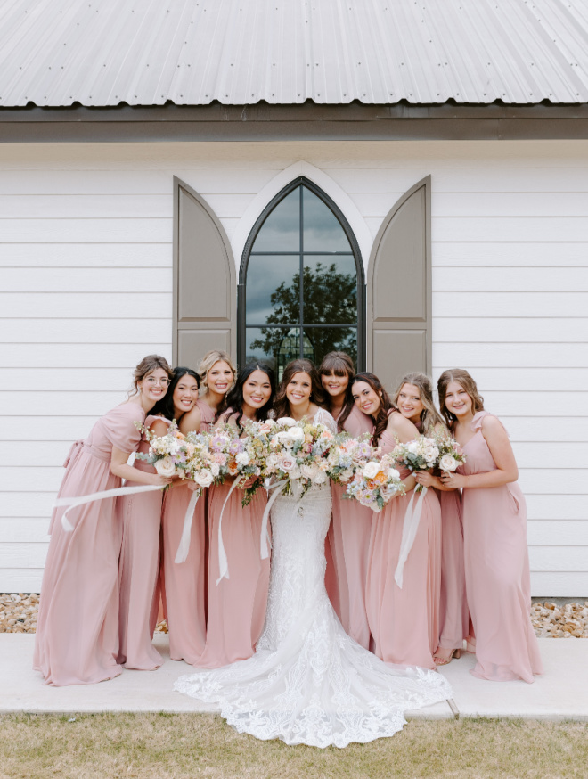 The bridesmaids in pastel pink dresses pose with the bride outside the hill country wedding venue, Deep in the Heart Farms.
