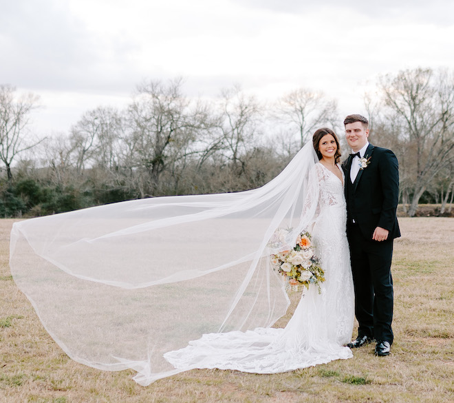 The bride and groom smiling in a field as the bride's veil flows in the wind.