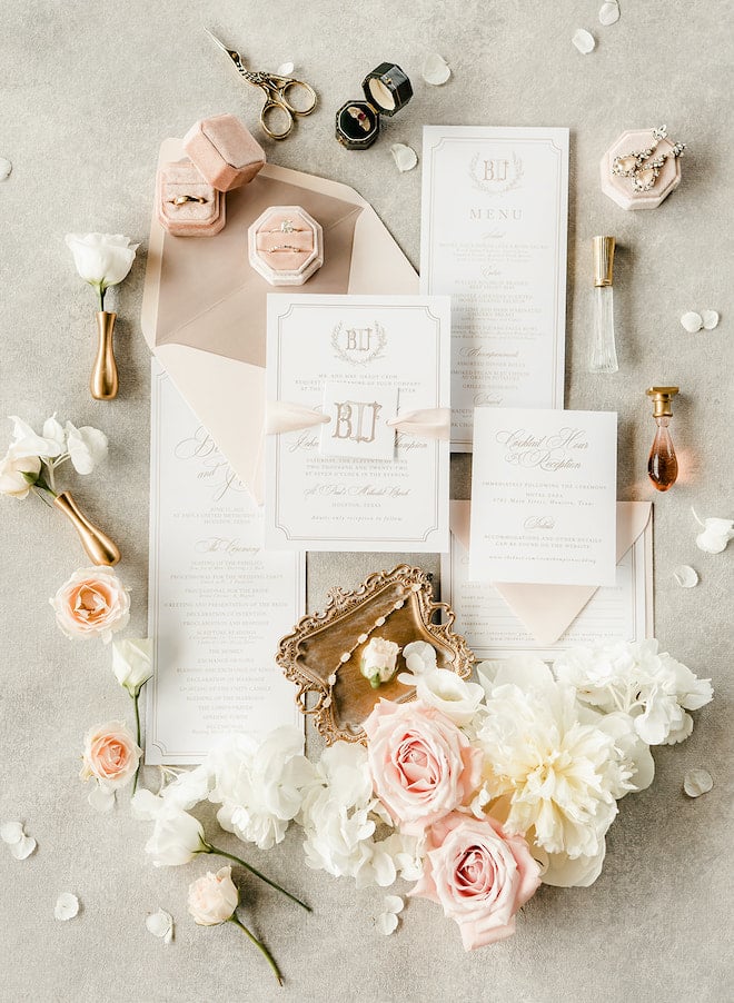 A flatlay of the wedding stationery invitations surrounded perfume, wedding bands and white and blush pink flowers. 