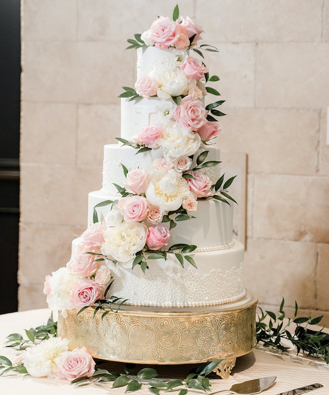 A 5-tier white wedding cake embellished with white, pink and green florals designed by Susie's Cakes sits on a gold cake stand. 