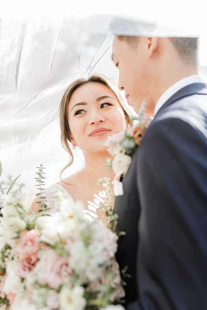 The groom looking at the bride as she smiles underneath her veil.