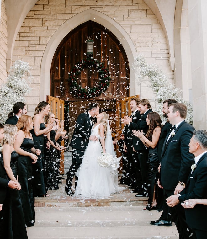 The bride and groom kissing in front of the church with the wedding party clapping.