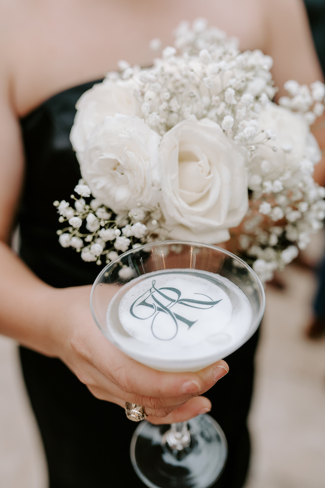 A bridesmaid holding her white bouquet and a drink with the couple's monogram on top.