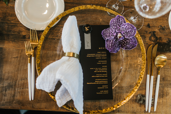 A black and gold menu on a clear charger with a gold rim, decorated with a purple flower.