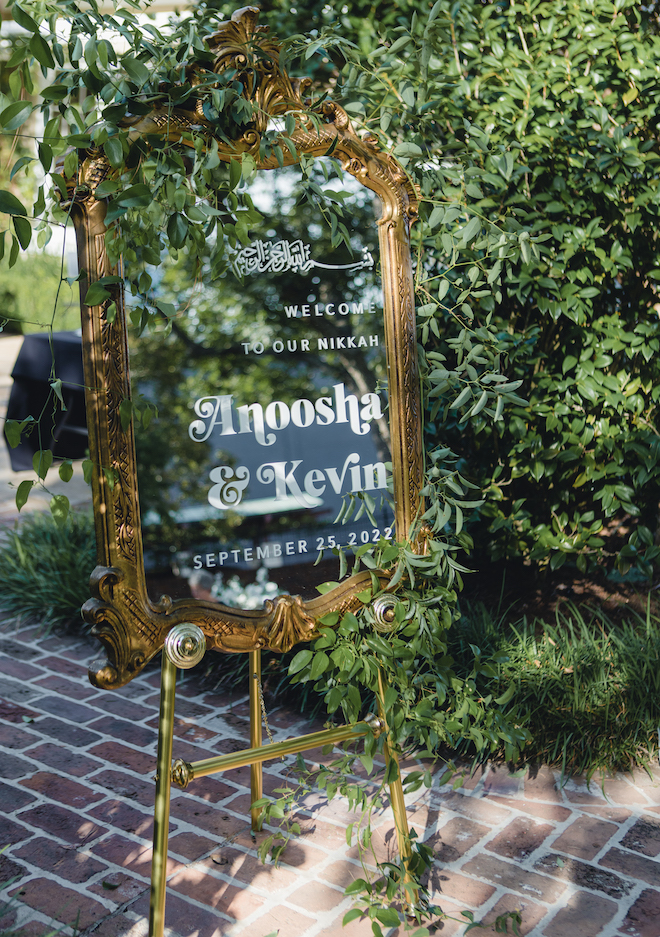 A clear sign with a gold frame that says "Welcome to our Nikkah. Anoosha & Kevin, September 25, 2022."