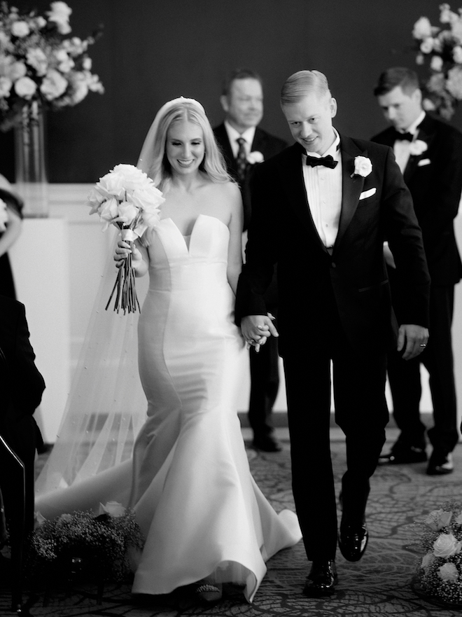 A black and white photo of the bride and groom holding hands as they walk down the aisle after their sophisticated ceremony at the Petroleum Club of Houston.
