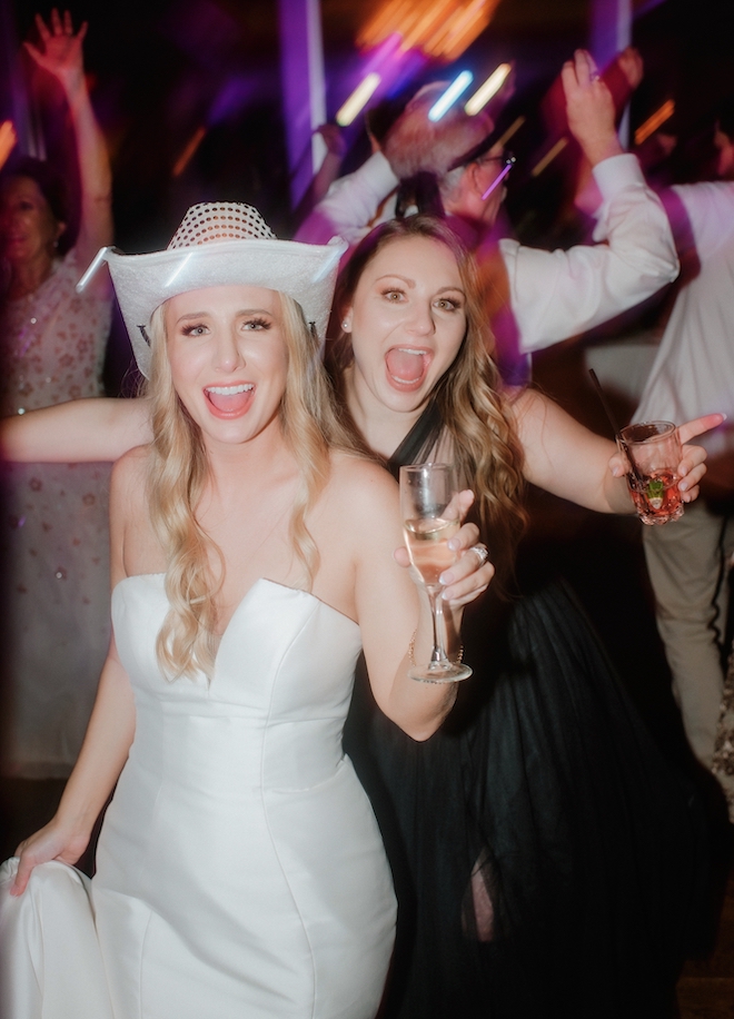 The bride in a white cowgirl hat and a bridesmaid smile as they are holding drinks on the dance floor.
