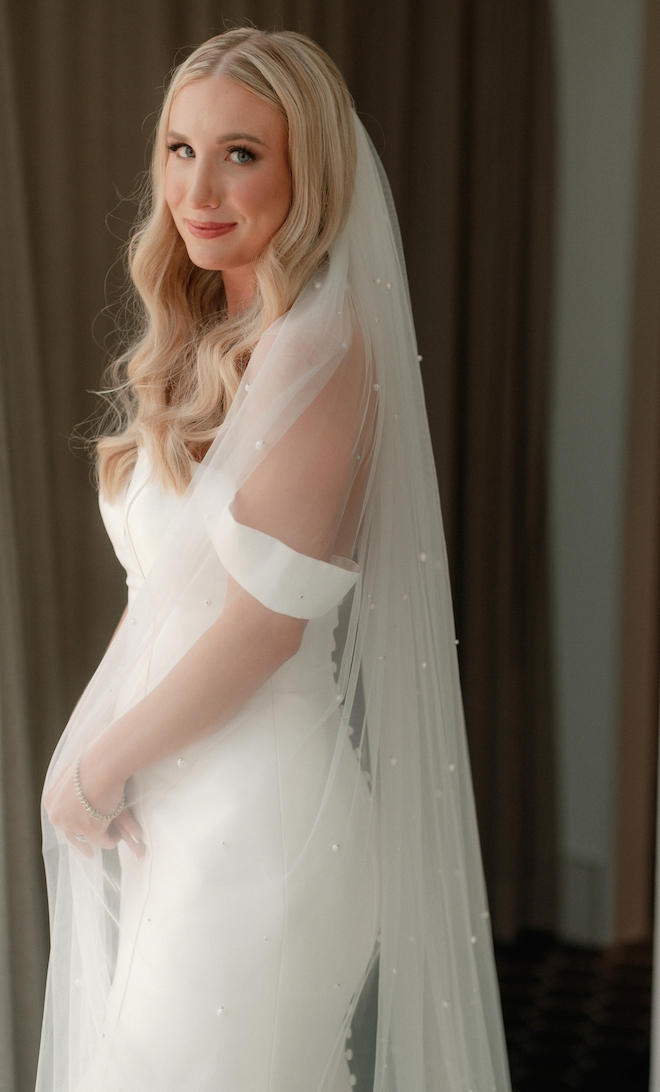 A blonde bride stands by a hotel room window on her wedding day in Houston