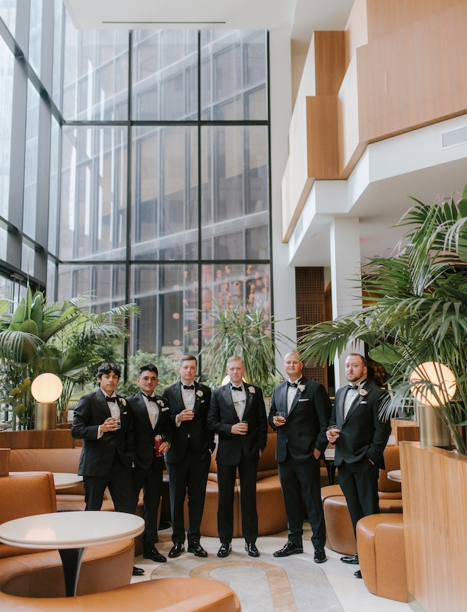 Groom stands with groomsmen holding cocktails in the bar lobby of downtown Houston hotel, C Baldwin