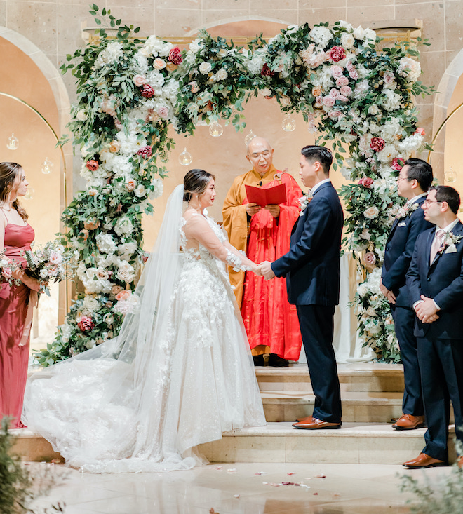 The bride and groom holding hands under a floral-filled altar at their wedding ceremony.