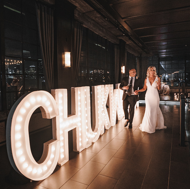 The bride and groom holding hands with lit-up marquee letters reading "Chukwuma."
