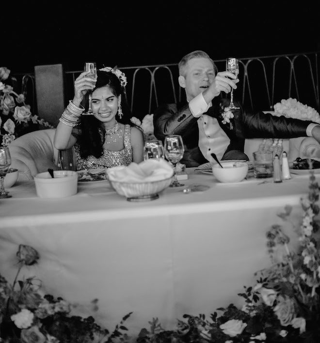 A black and white photo of the bride and groom raising a glass of champagne at their wedding reception table. 