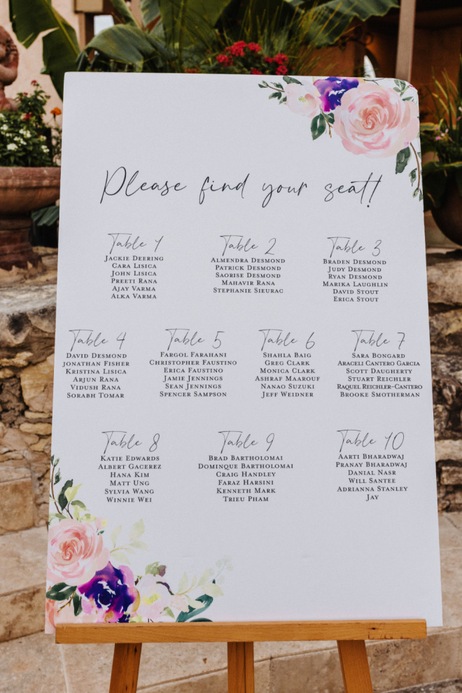 A white and pastel seating chart displayed on an easel at the outdoor wedding reception. 