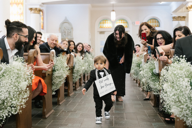A little boy walking down the aisle with a sign that reads "Get Ready Amo George, here she comes"