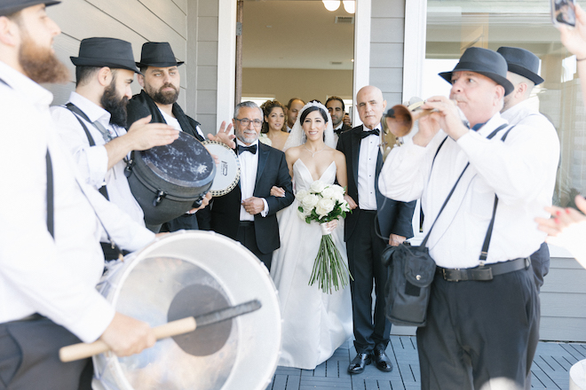 The groom and her father walking out of the house while men play instruments.