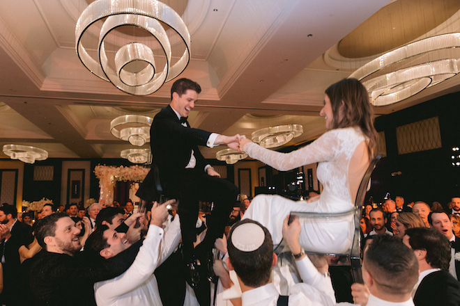 The bride and groom holding hands as they are lifted into the air on their chairs.