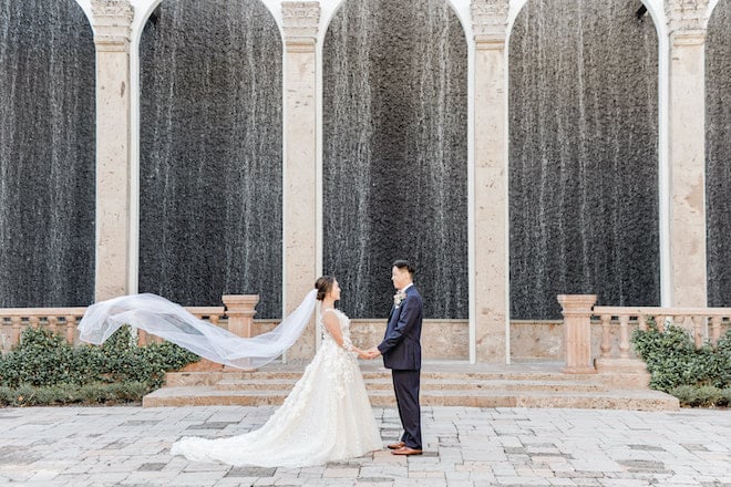 The brides veil flowing in the wind as they hold hands in front of the water wall at The Bell Tower on 34th.