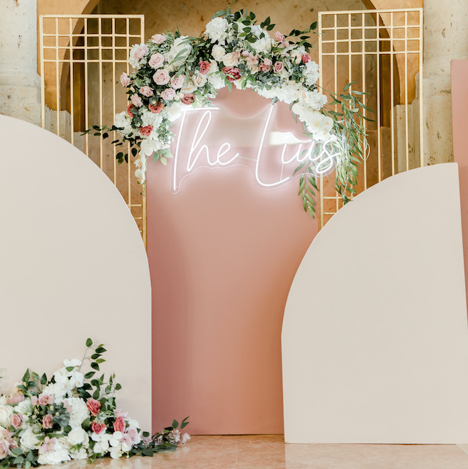 A lit up sign reading "The Lius" on a pink background and decorated in florals.