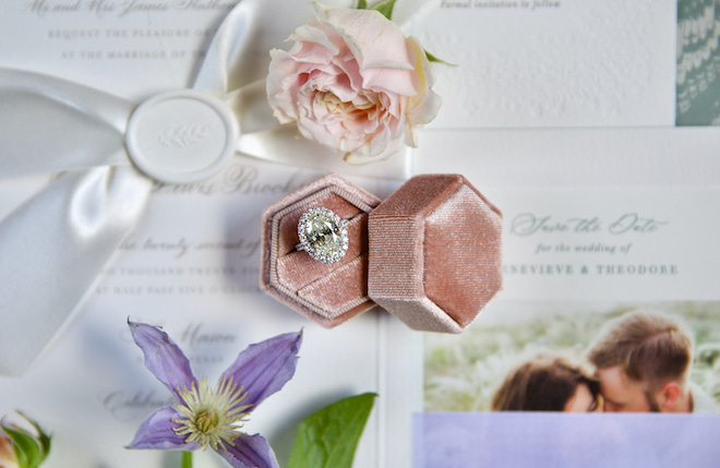 A flatlay of the white wedding stationery with roses and an oval-shaped diamond in a pink velvet ring box. 