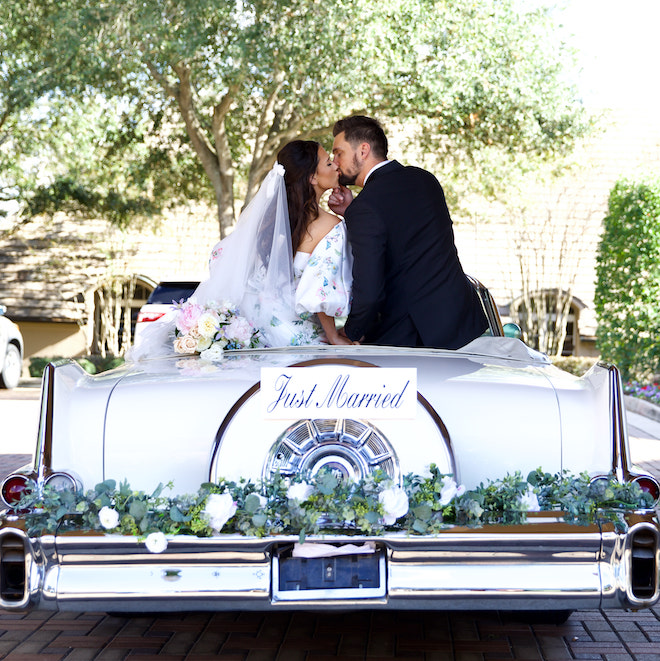 The bride and groom kissing on a vintage car with a "just married" sign and flowers on the bumper.