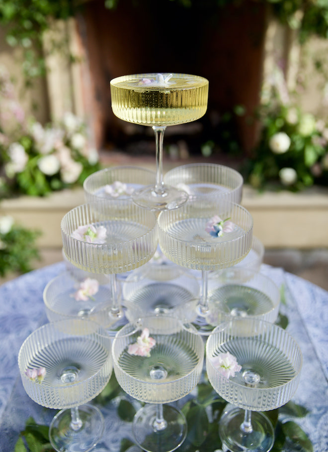 Circular cocktail glasses are stacked into a champagne tower and garnished with pink flower petals, adding a playful feel.