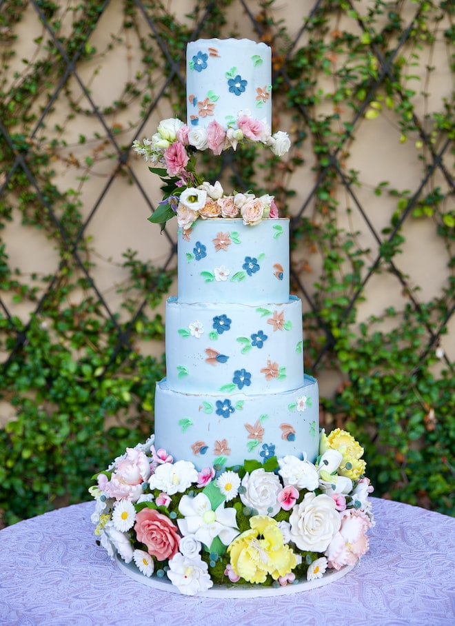 A playful four-tier light blue wedding cake is detailed in various pastel colored florals. 