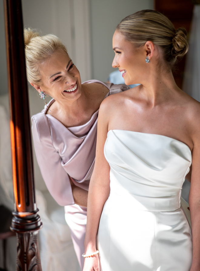 The mother of the bride helping the bride with her dress as they smile.