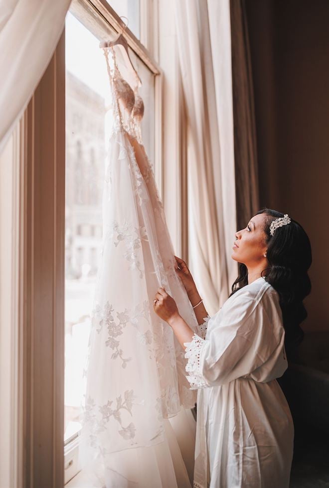 A bride wearing a robe gazes at her galia lahav gown hung up on a windowsill at houston wedding venue and hotel, Hotel Icon, on her wedding day at The Corinthian Houston. 