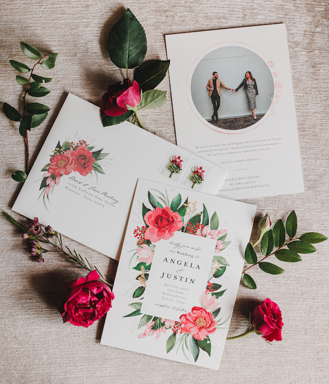 A wedding invitation suite for Angela and Justin with a photo of them holding hands and pink flowers and greenery on the invitations. 