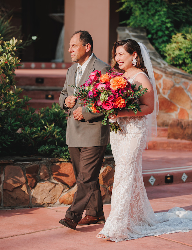 The bride smiling walking down the aisle with her father. She is holding a large bouquet of vibrant pink and orange florals. 