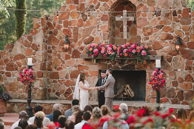 The bride and groom holding hands at their alfresco ceremony. The altar is decorated with pink and orange florals.