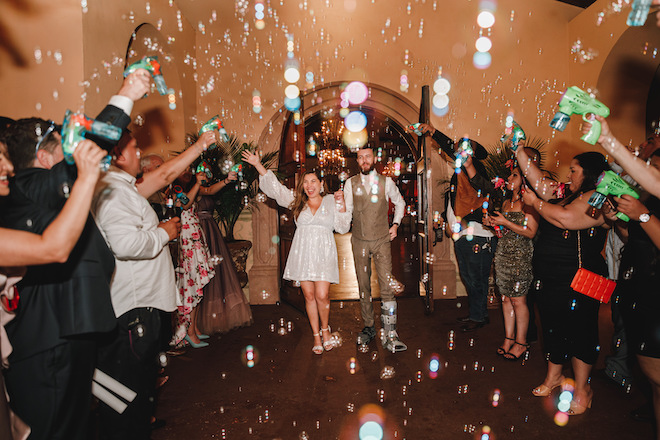The bride and groom holding hands during their bubble sendoff while guests cheer on either side of them holding bubble guns. 