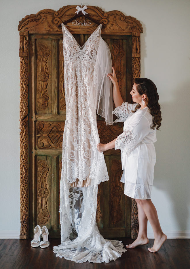 The bride wearing a white robe looking at her lace wedding gown hung up on a brown door. 
