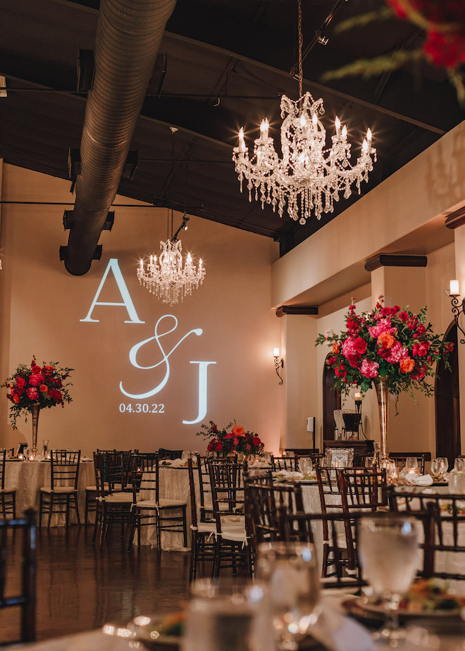 The reception space decorated with vibrant pink and orange floral centerpieces, chandeliers and an "A&J 4.30.22" on the wall.