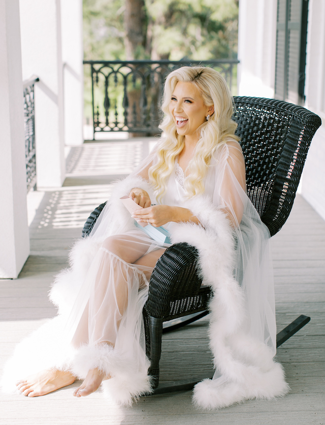 The bride sitting in a black wicker rocking chair on a white porch smiling while holding a letter. She is wearing a white sheer feather robe.