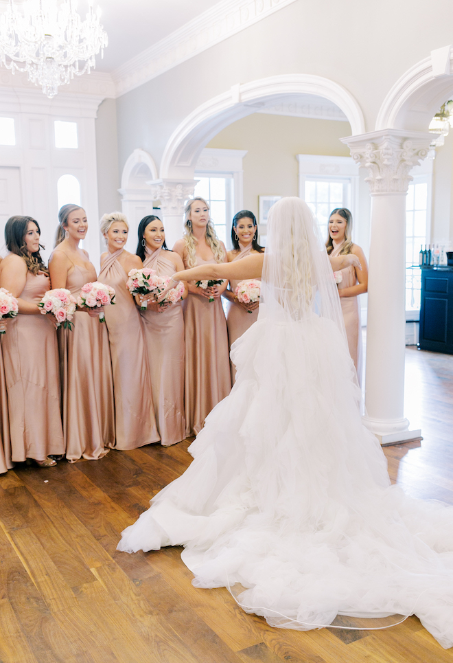 The back of the brides dress and veil with her arms out as her bridesmaids smile at her.