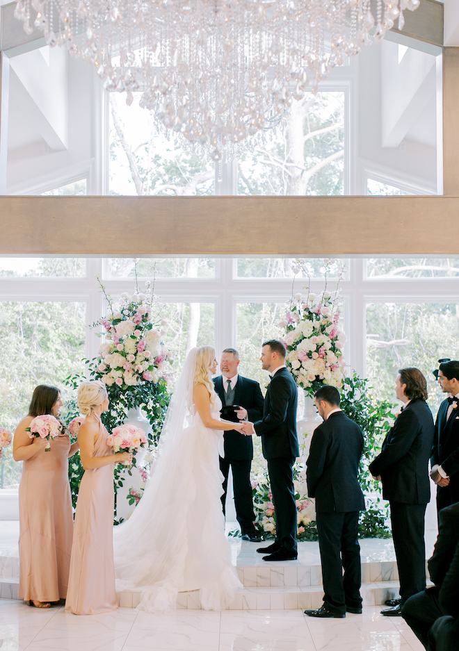 The bride and groom holding hands at the altar with greenery and light pink and ivory florals. The bridesmaids and groomsmen stand on either side of her. 