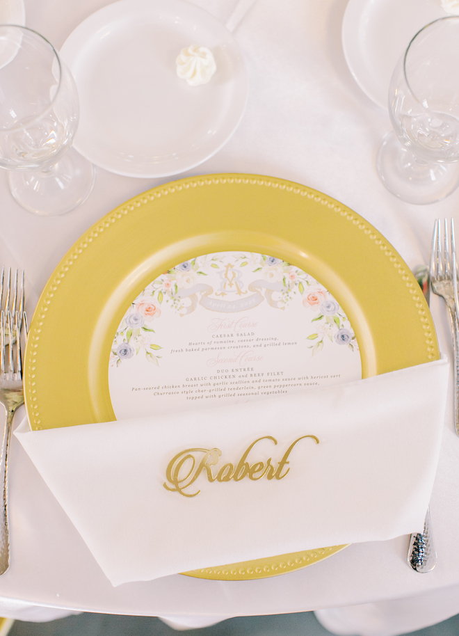 A yellow gold plate with a floral-print light and bright menu and a white napkin with "Robert" written on it in gold. 