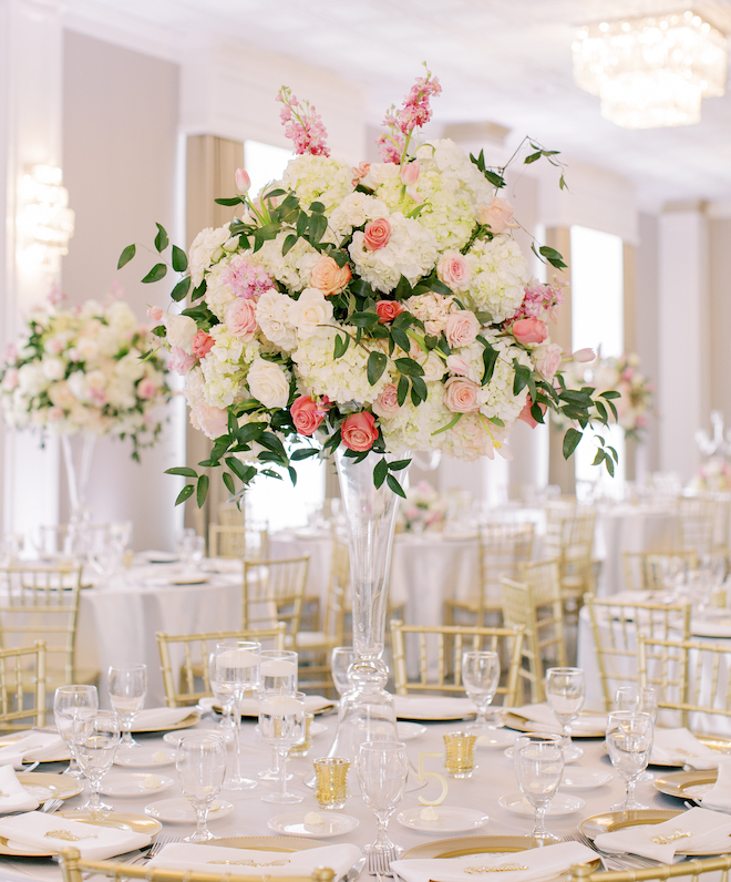 A floral centerpiece with white and pink florals in a tall glass vase on a round reception table with gold accents. 