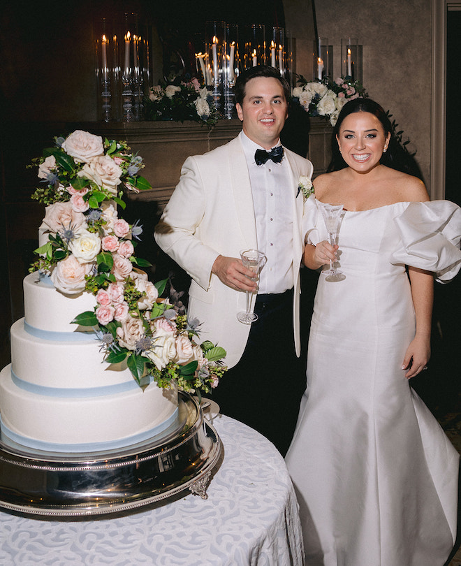 The couple smiling with champagne glasses next to their light blue, pink and cream wedding cake.