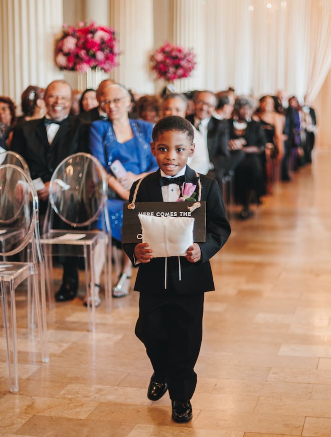 A boy walking down the aisle with a "here comes the bride" sign and a pillow for the bride and groom's wedding bands during a ceremony at the corinthian in downtown houston