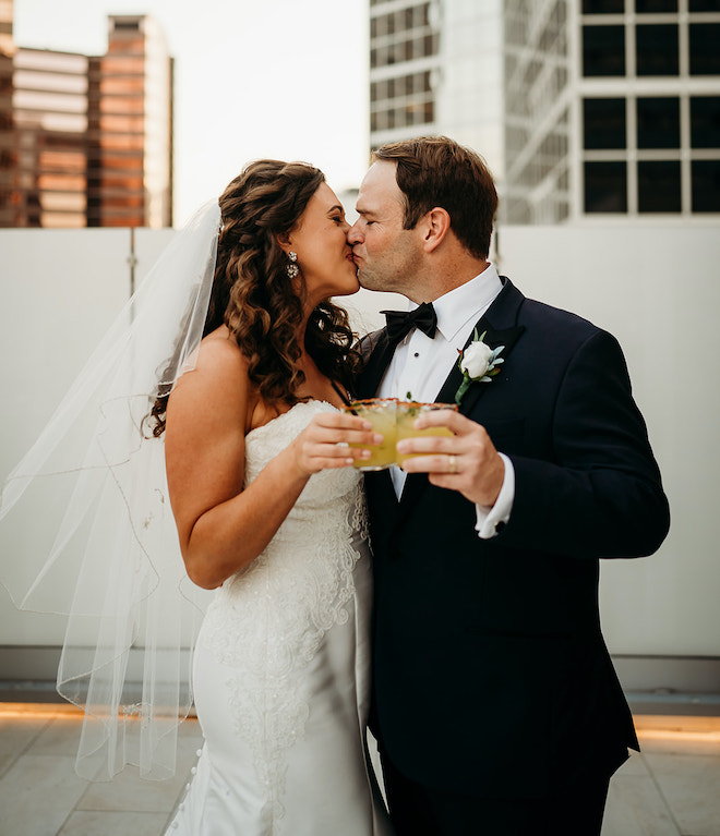 The bride and groom kissing as they clink their cocktails at their rooftop wedding.